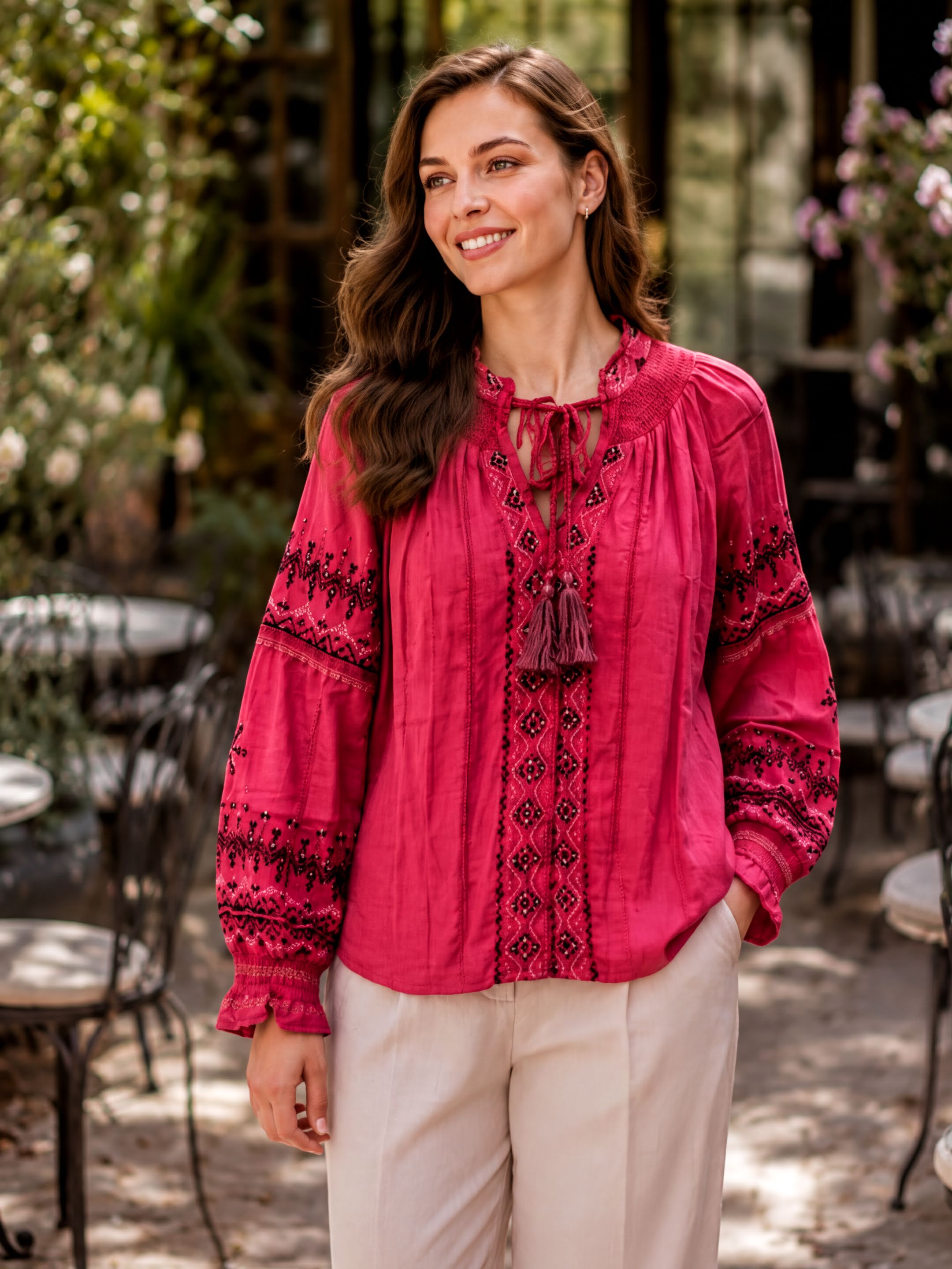 A woman wearing a bright pink blouse with embroidered patterns and a tassel, standing in an outdoor setting with a table and chairs visible in the background.