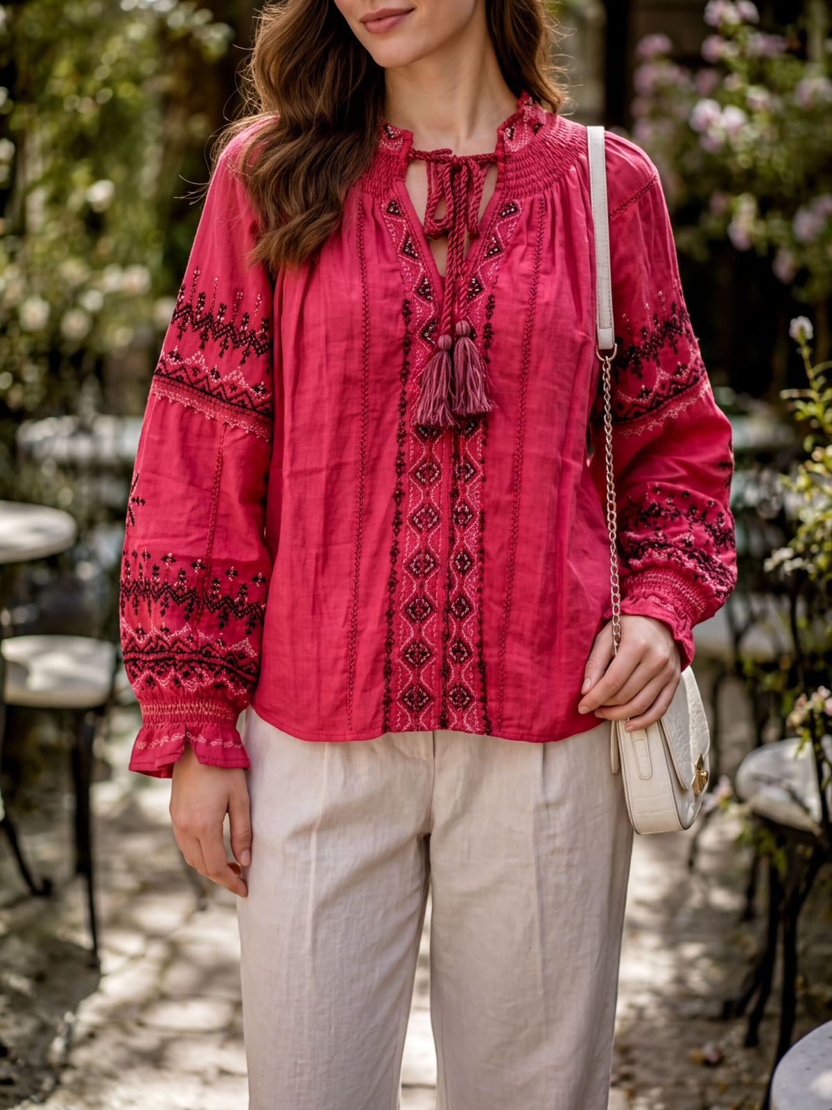 A woman wearing a bright red blouse with intricate embroidery and a white purse stands in a garden setting.