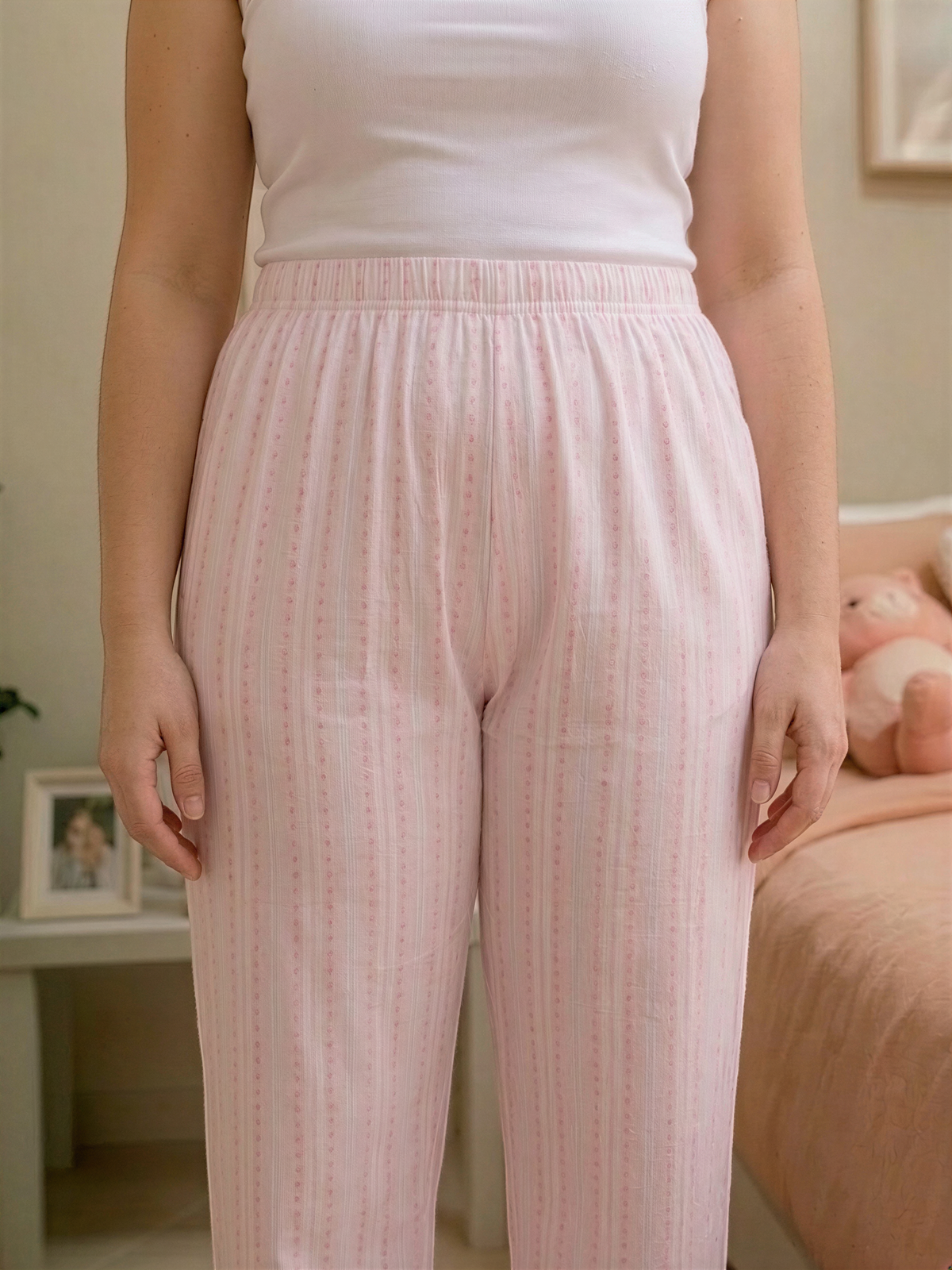 A woman wearing pink striped pajama pants stands in a room with a bed and a framed photo visible in the background.