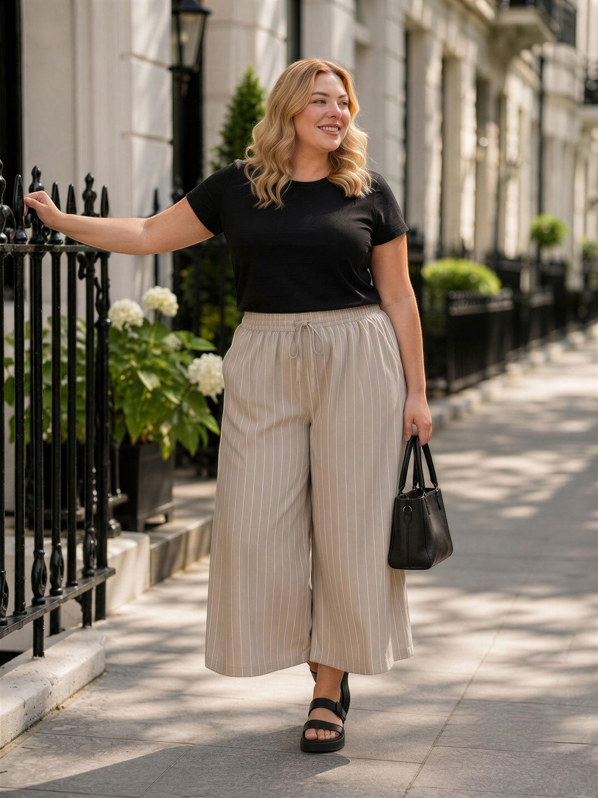 A woman in a black top and beige striped pants is walking down a sidewalk, leaning on a black fence and carrying a black handbag.