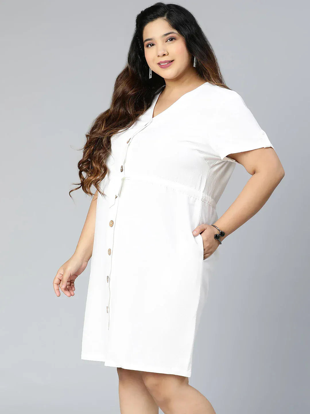 A woman wearing a white dress with buttons and holding a purse is standing against a white background.