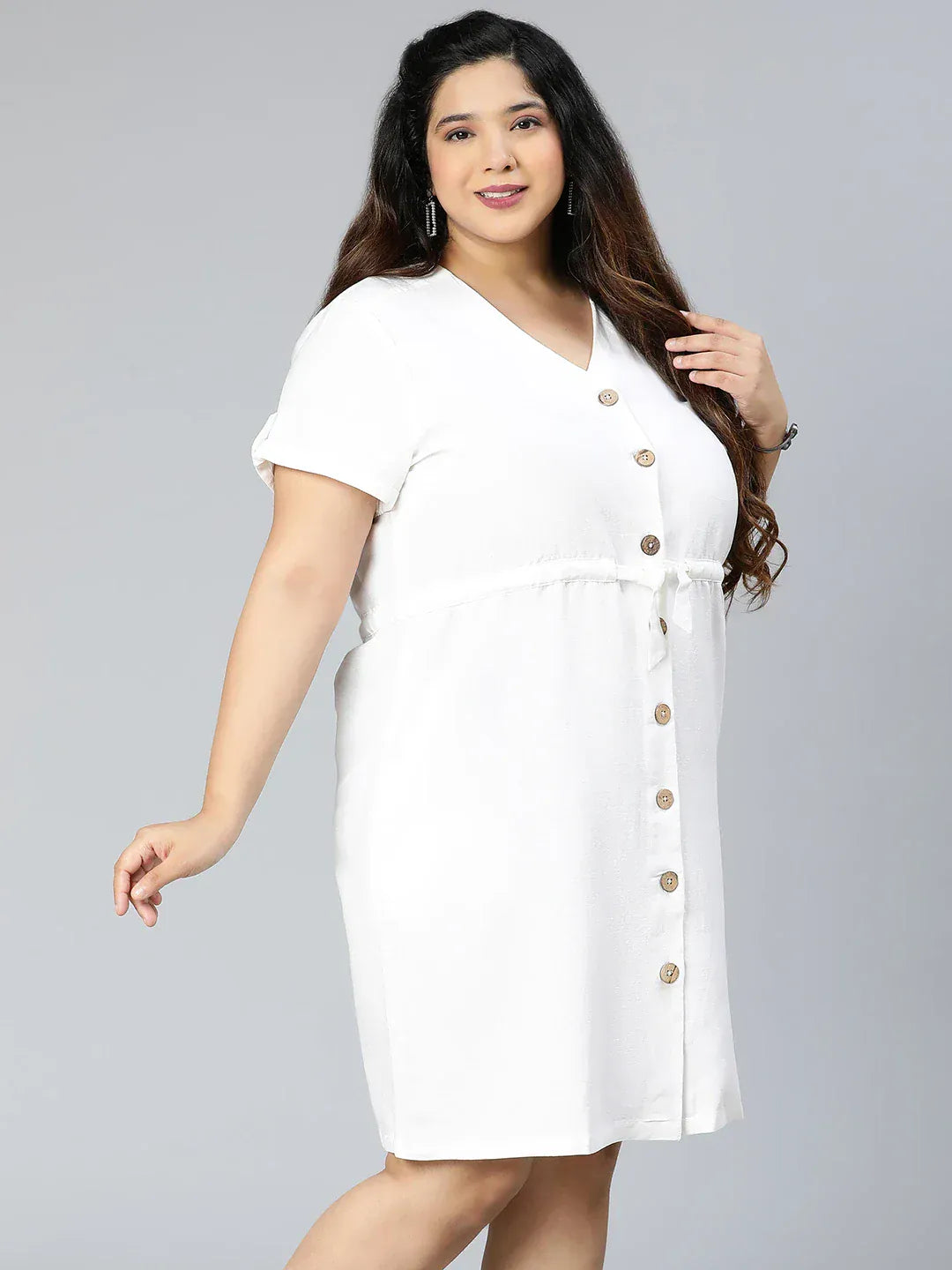 A woman wearing a white dress with buttons and holding a purse is standing against a white background.