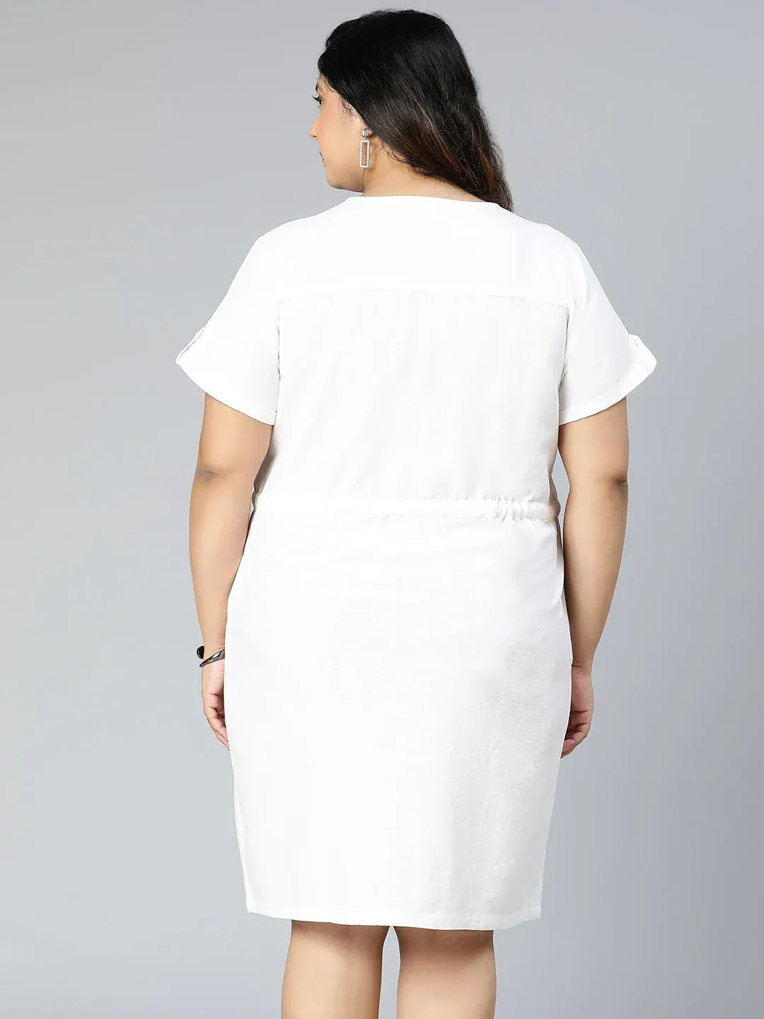 A woman wearing a white dress with buttons and holding a purse is standing against a white background.