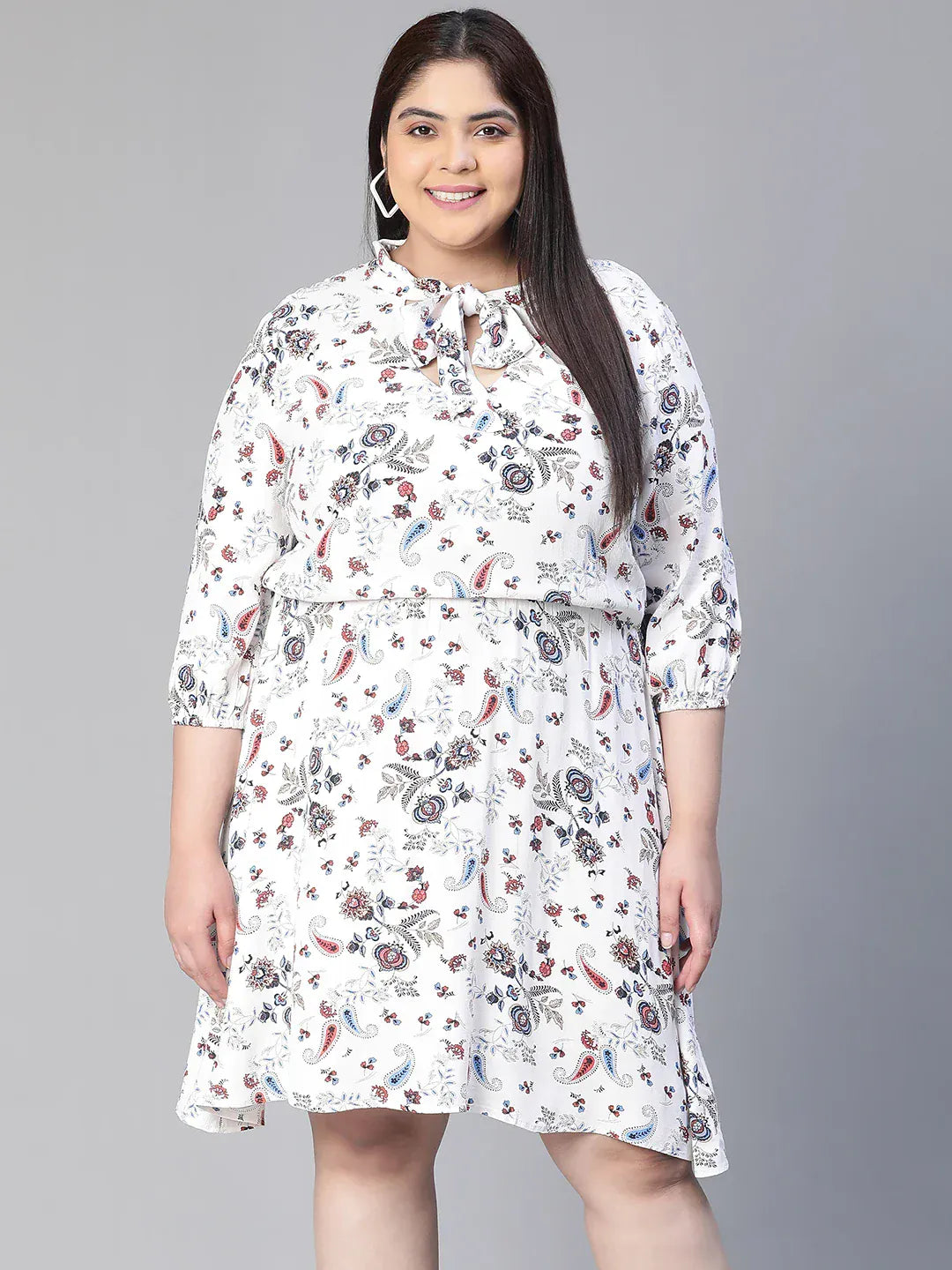 A woman wearing a white dress with a floral print, smiling and holding a small white purse.