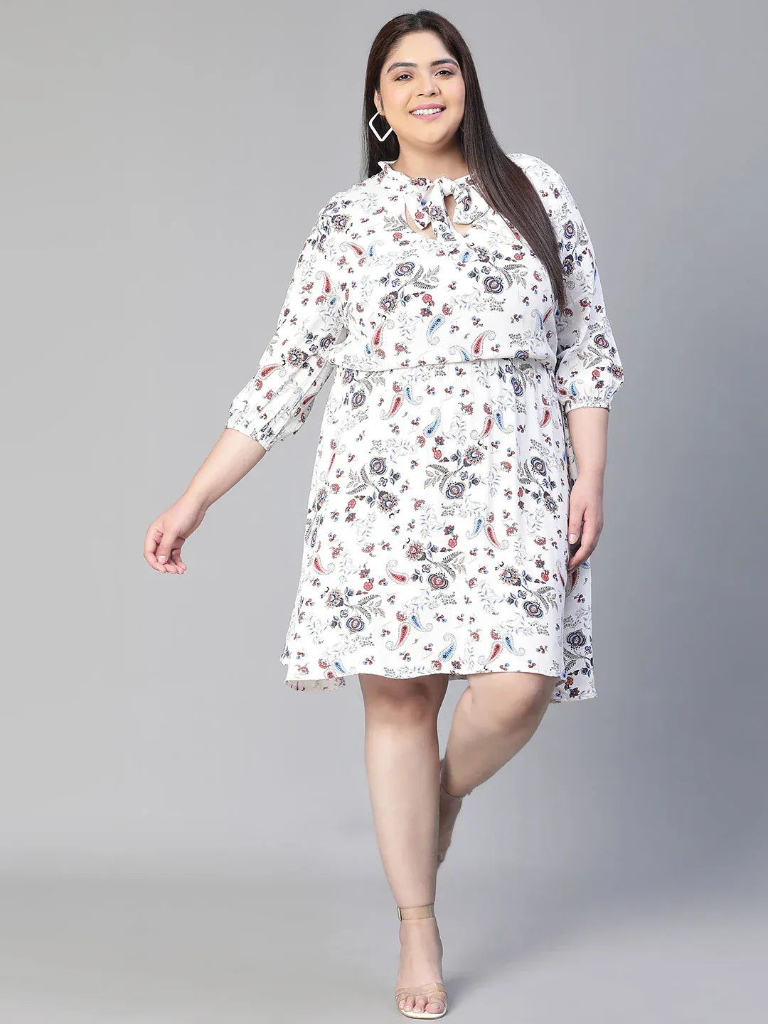 A woman wearing a white dress with a floral print, smiling and holding a small white purse.