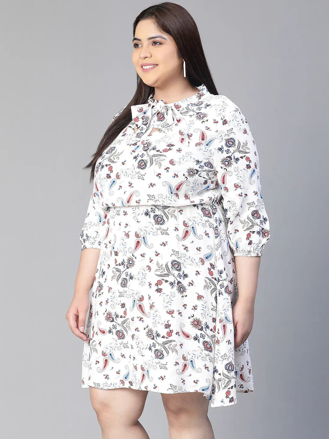 A woman wearing a white dress with a floral print, smiling and holding a small white purse.