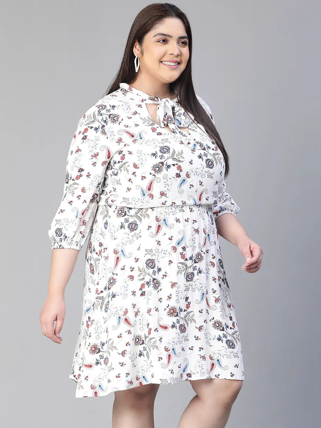 A woman wearing a white dress with a floral print, smiling and holding a small white purse.