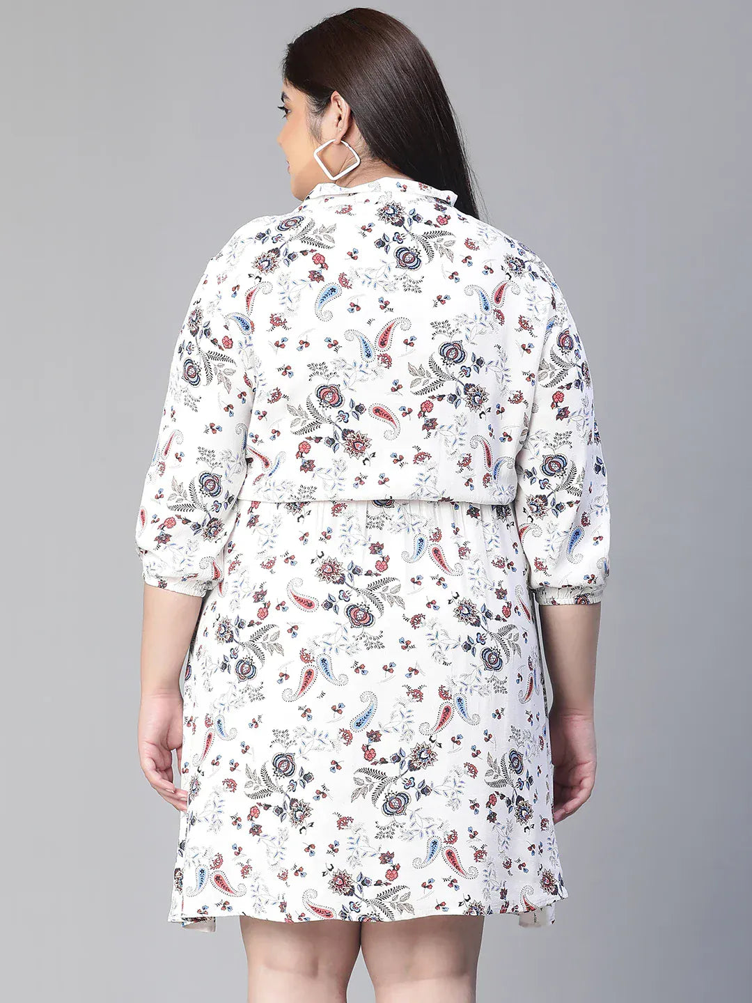 A woman wearing a white dress with a floral print, smiling and holding a small white purse.