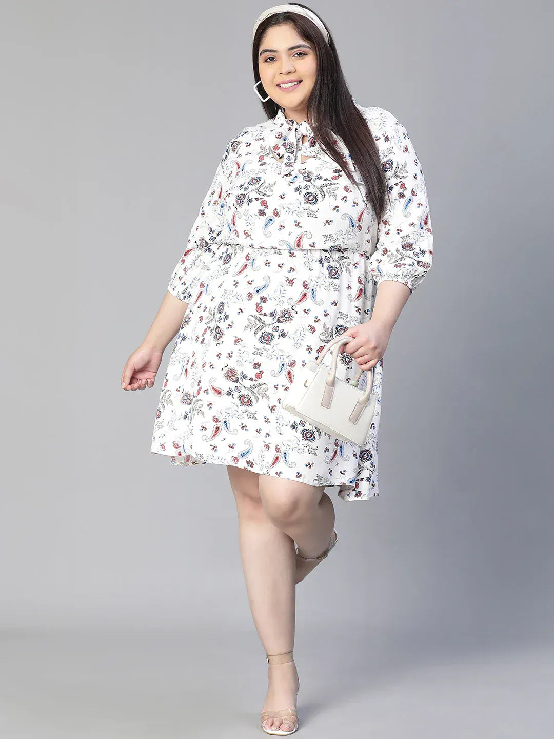 A woman wearing a white dress with a floral print, smiling and holding a small white purse.