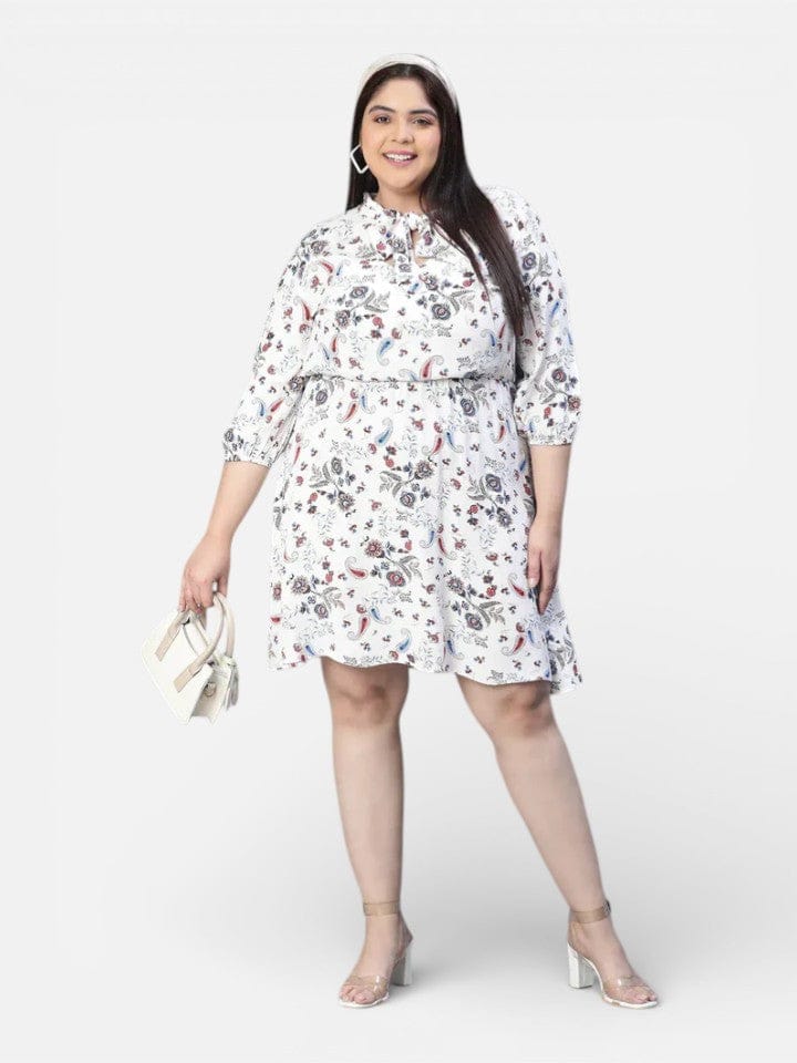 A woman wearing a white dress with a floral print, smiling and holding a small white purse.