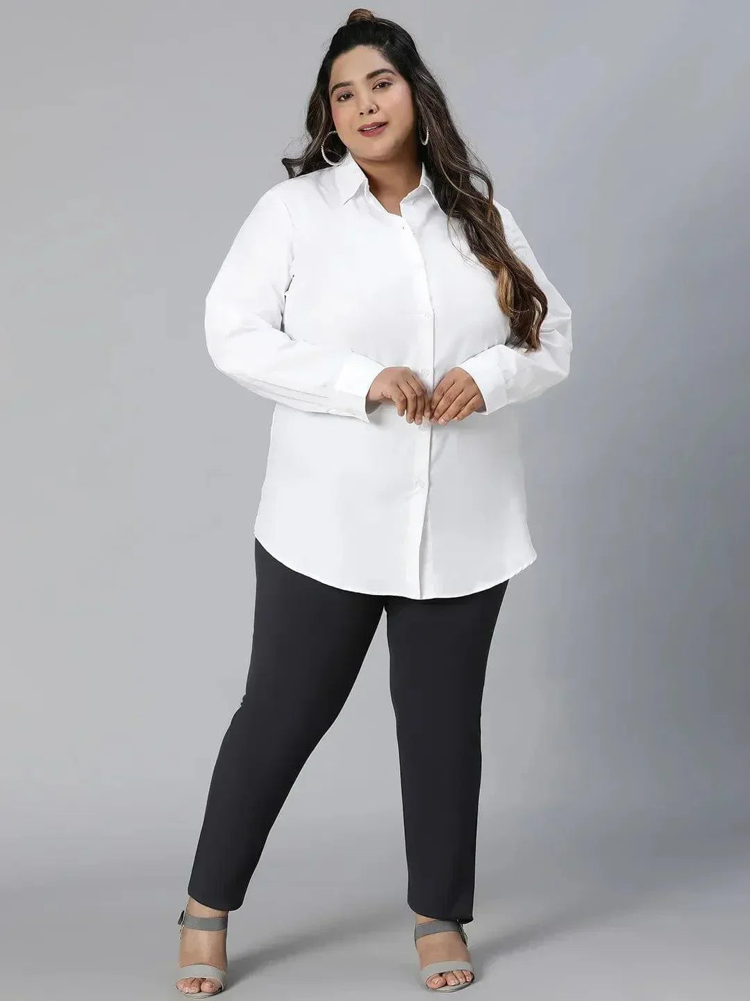A woman with long dark hair wearing a white button-up shirt and black pants is smiling at the camera.