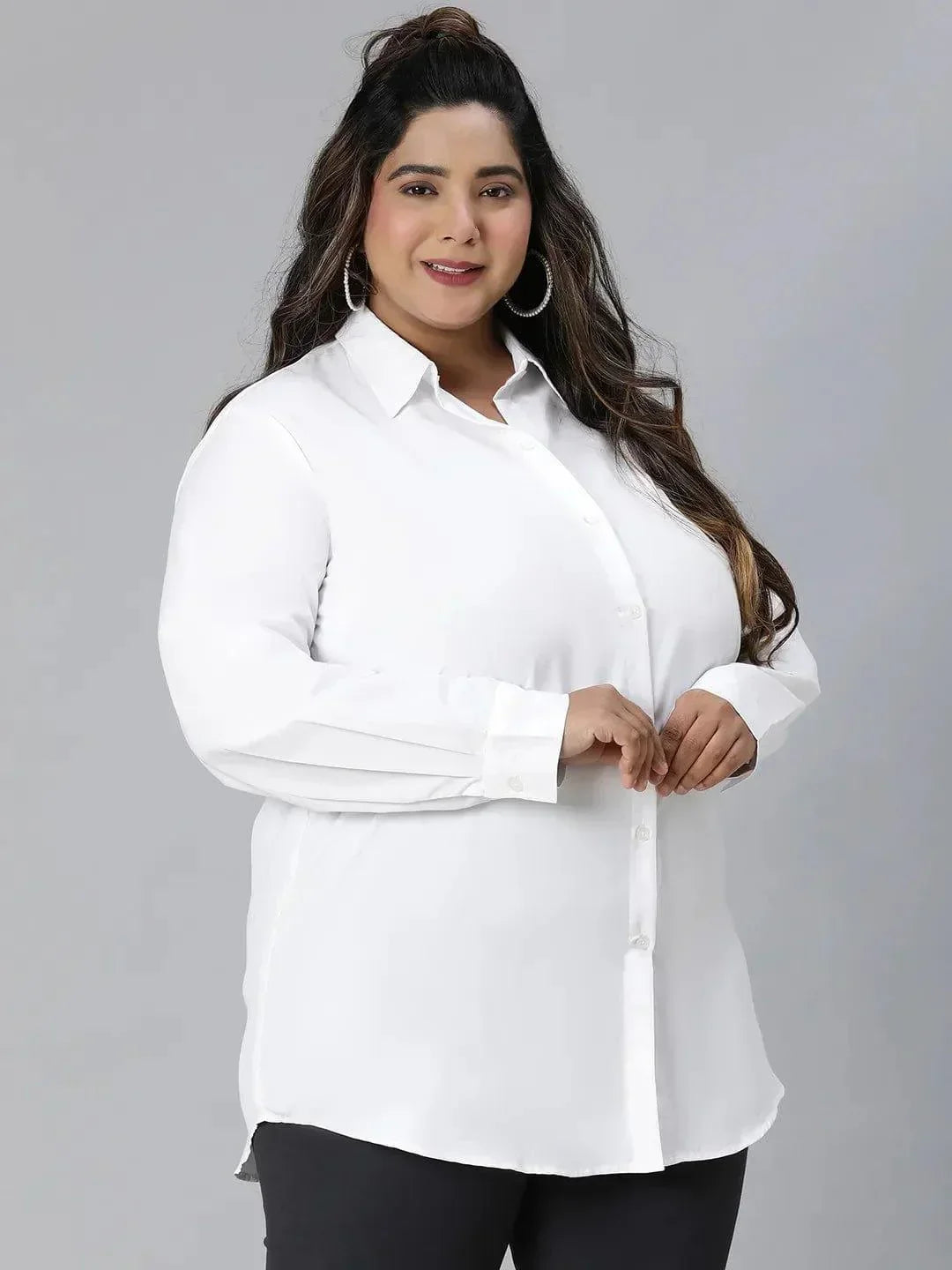 A woman with long dark hair wearing a white button-up shirt and black pants is smiling at the camera.
