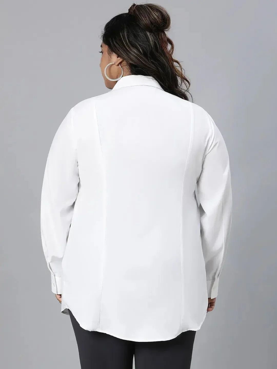 A woman with long dark hair wearing a white button-up shirt and black pants is smiling at the camera.
