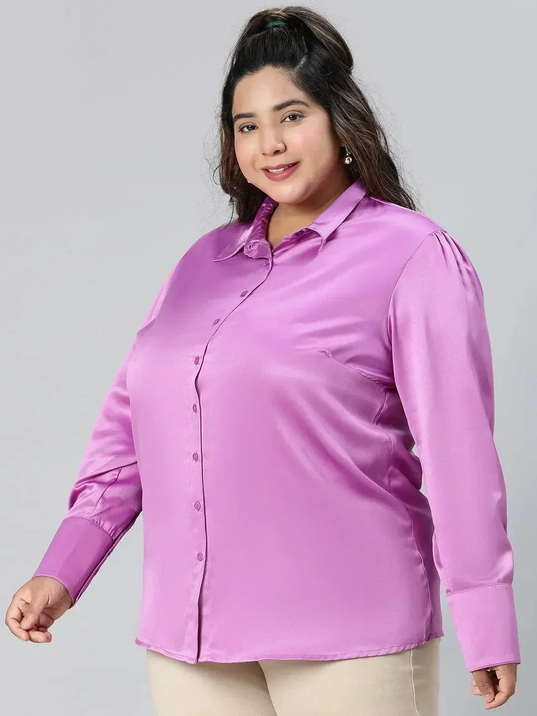 A woman wearing a pink shirt and beige pants is posing with her hand on her head and smiling.
