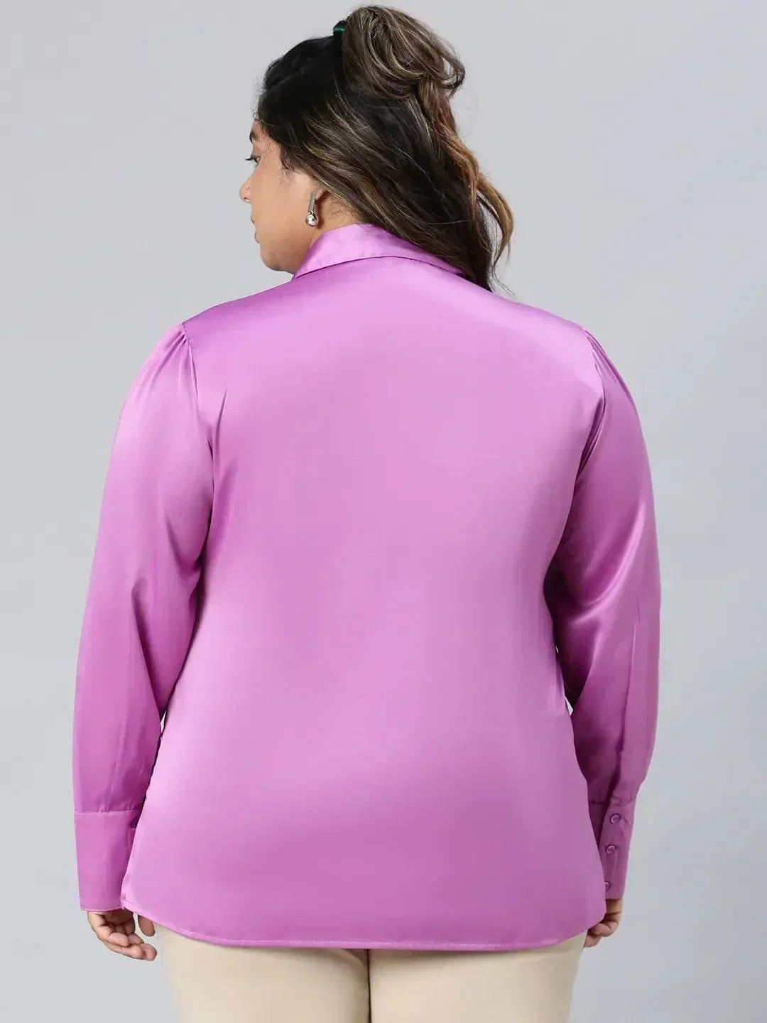 A woman wearing a pink shirt and beige pants is posing with her hand on her head and smiling.