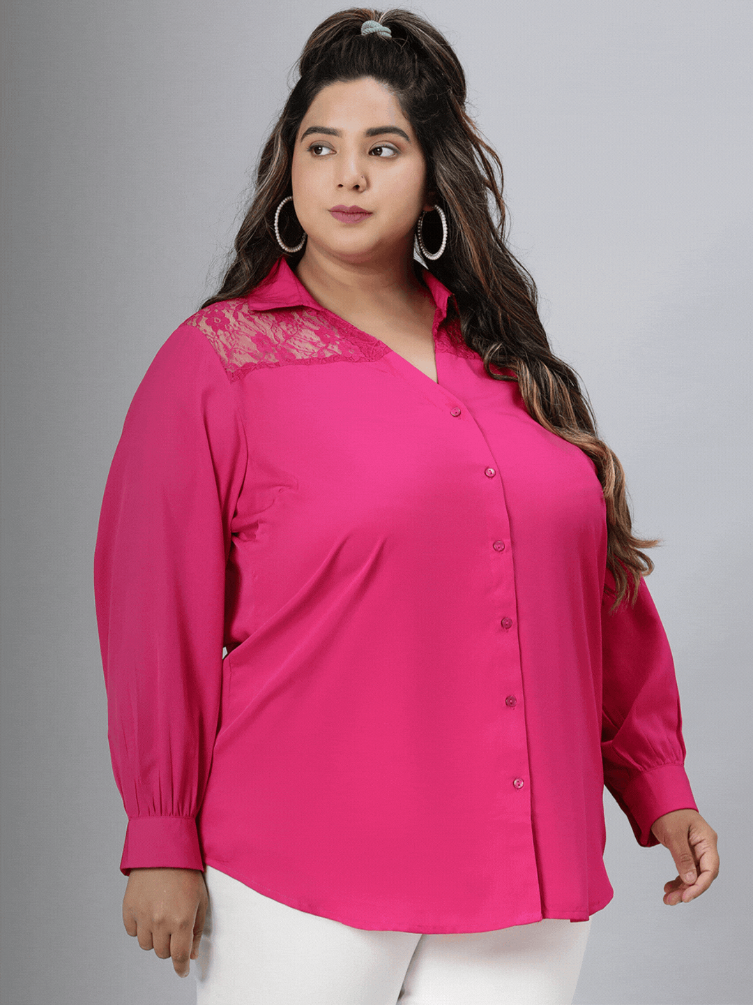 A woman wearing a bright pink blouse, white pants, and sunglasses, holding a small purse, standing against a white background.