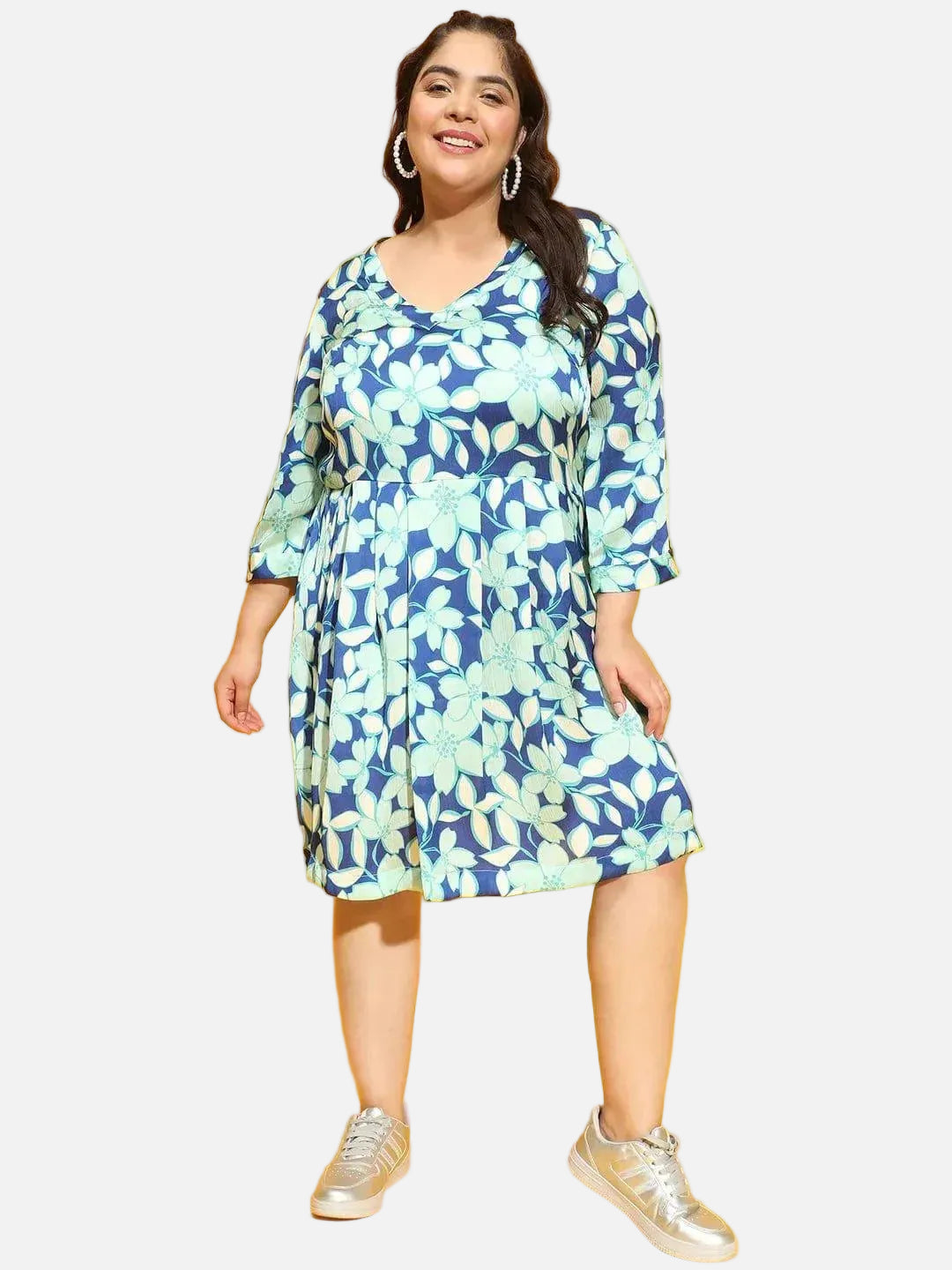 A woman wearing a blue and green floral print dress and silver sneakers is smiling at the camera.