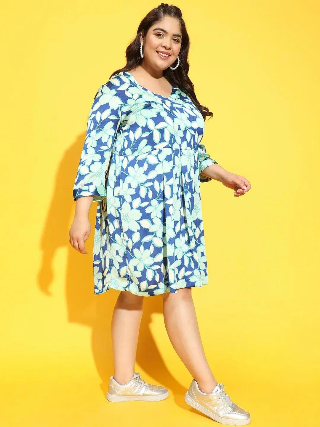 A woman wearing a blue and green floral print dress and silver sneakers is smiling at the camera.