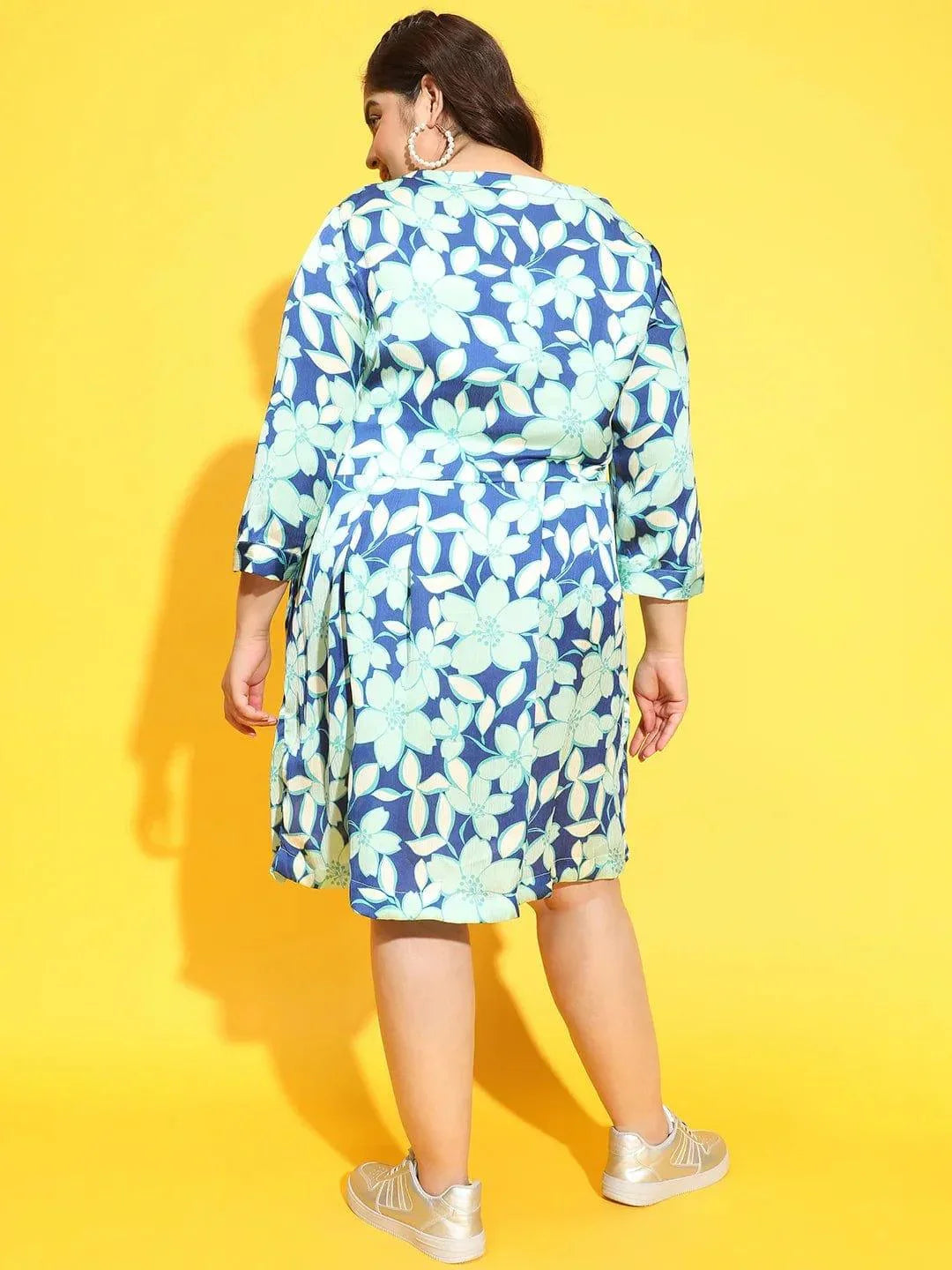 A woman wearing a blue and green floral print dress and silver sneakers is smiling at the camera.