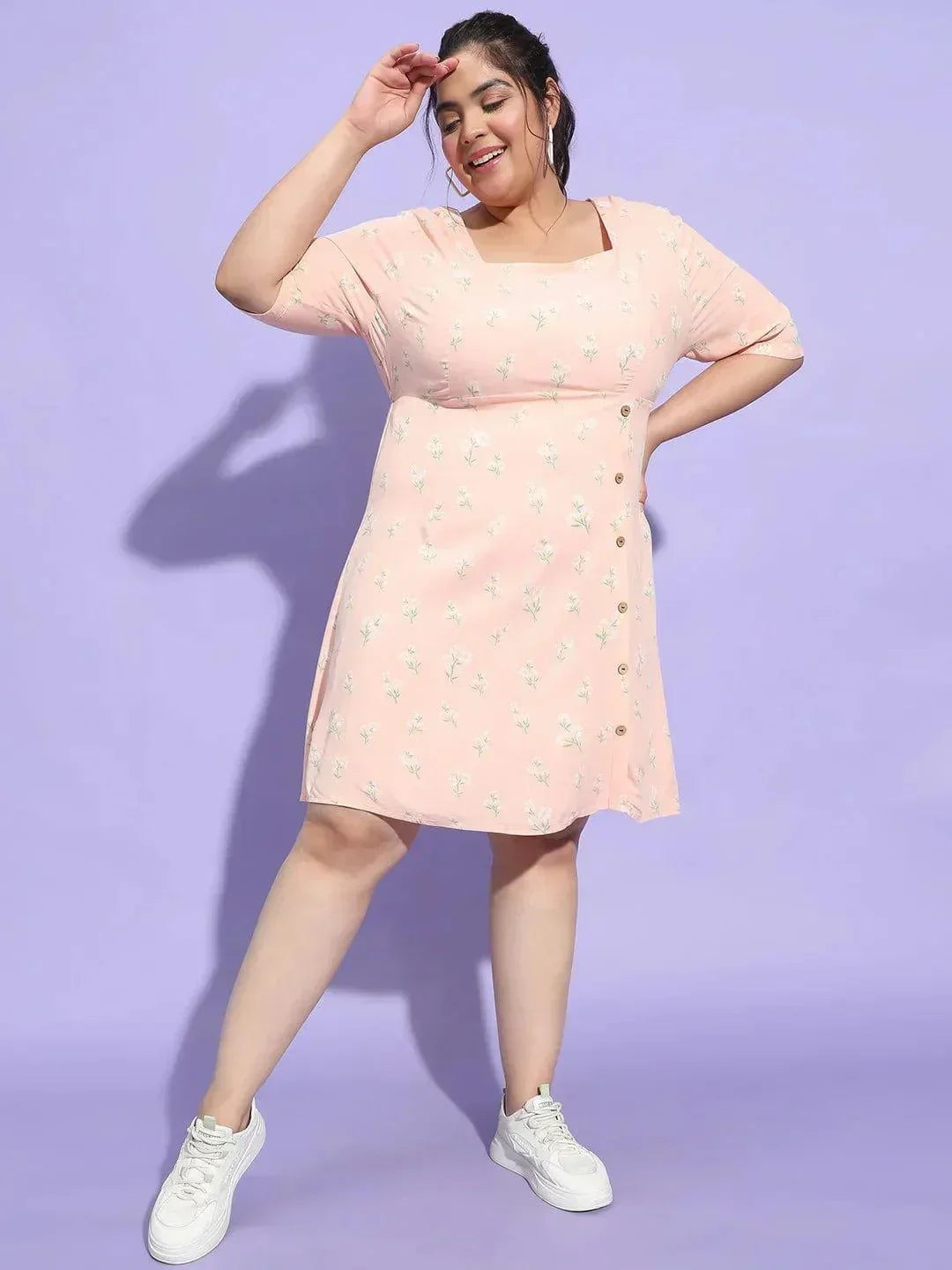 A woman wearing a pink dress with a floral pattern and white sneakers is smiling and posing for the camera.