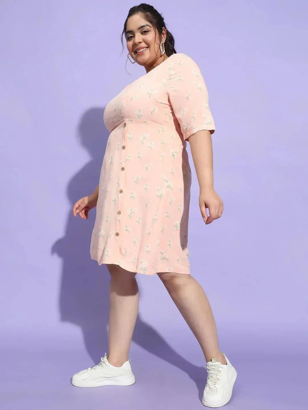 A woman wearing a pink dress with a floral pattern and white sneakers is smiling and posing for the camera.