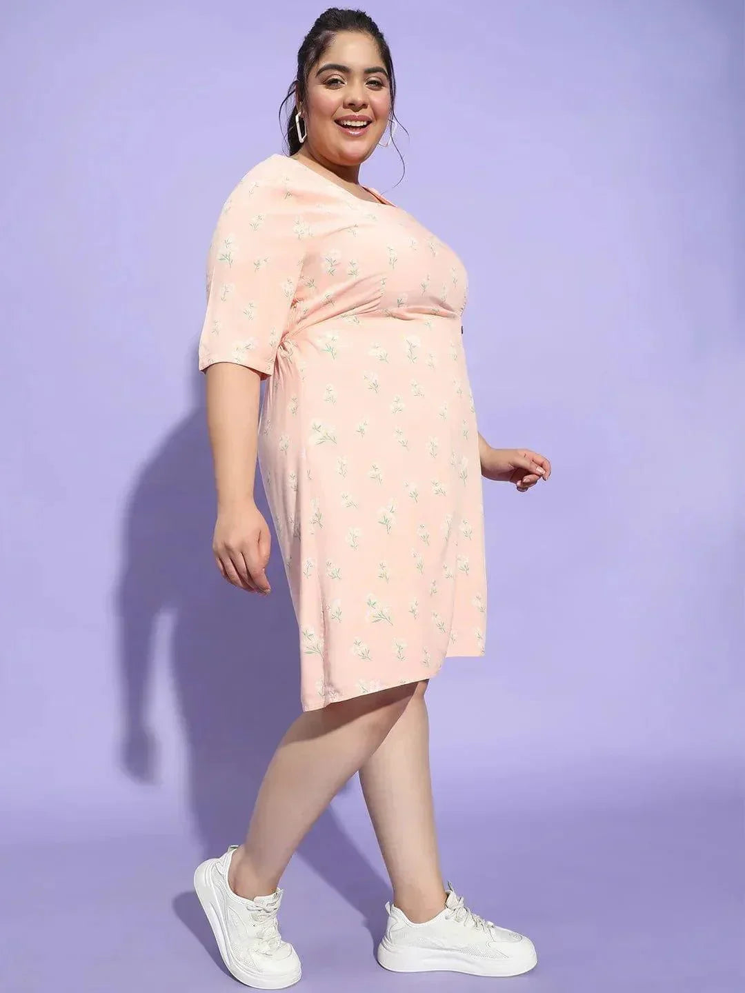 A woman wearing a pink dress with a floral pattern and white sneakers is smiling and posing for the camera.