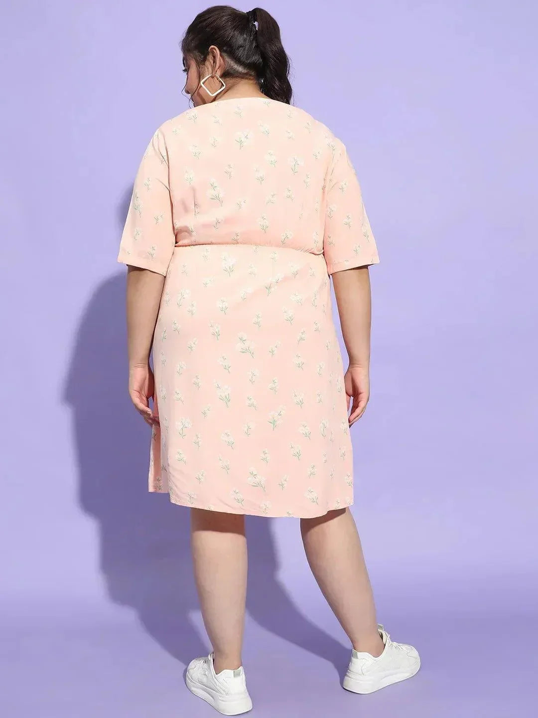 A woman wearing a pink dress with a floral pattern and white sneakers is smiling and posing for the camera.