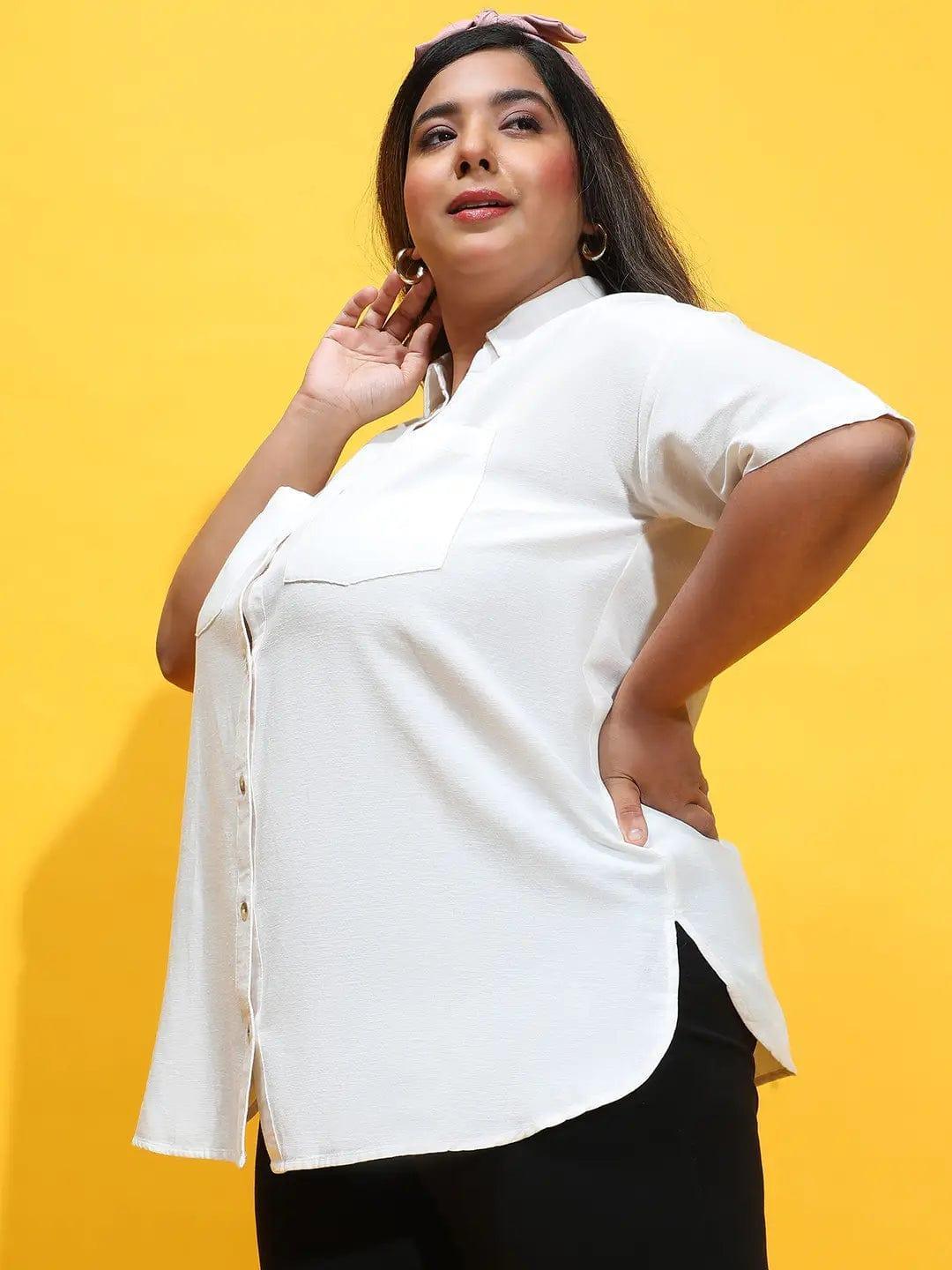 A woman wearing a white shirt and black pants is posing with her hands in her hair.
