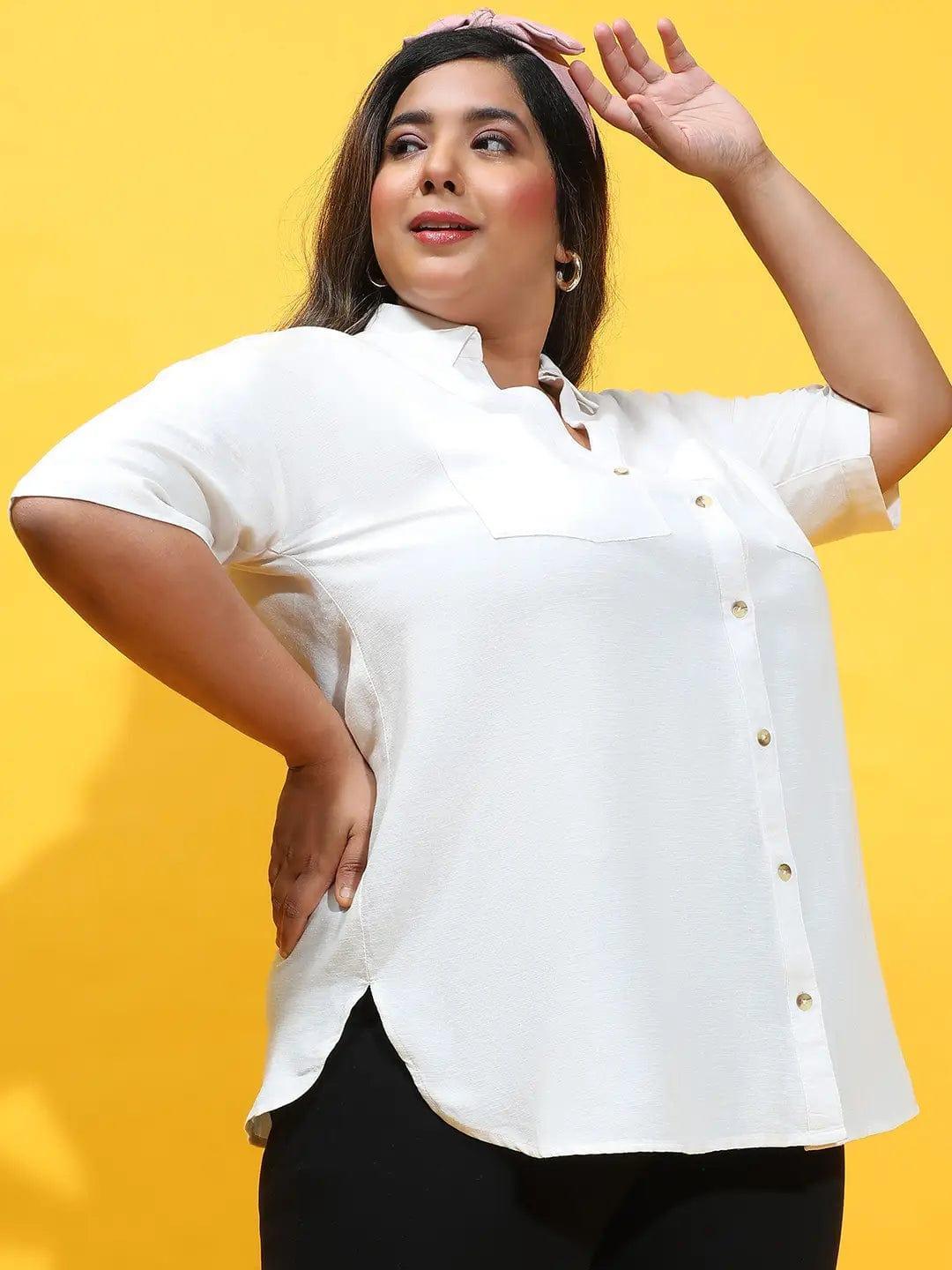 A woman wearing a white shirt and black pants is posing with her hands in her hair.
