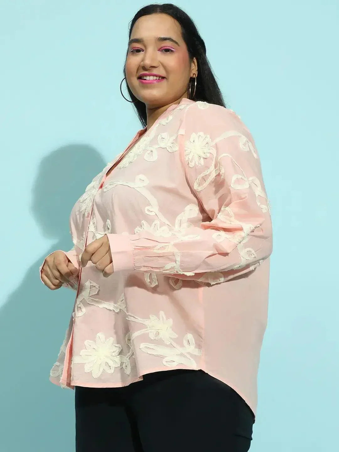 A woman with long dark hair wearing a pink blouse with white floral embroidery.