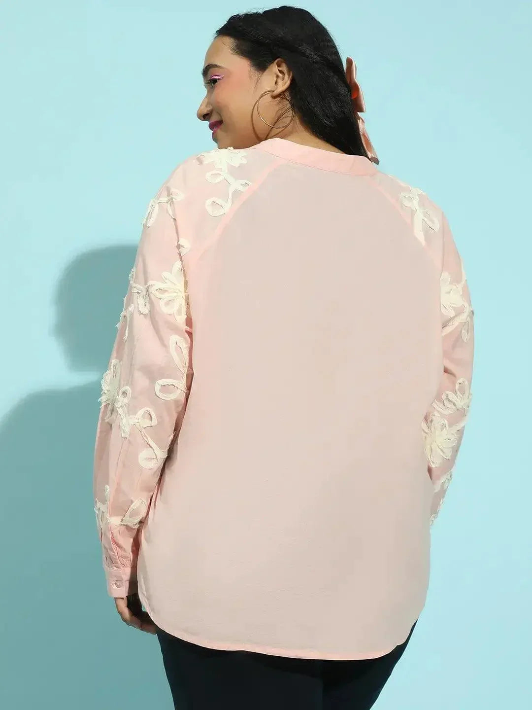 A woman with long dark hair wearing a pink blouse with white floral embroidery.