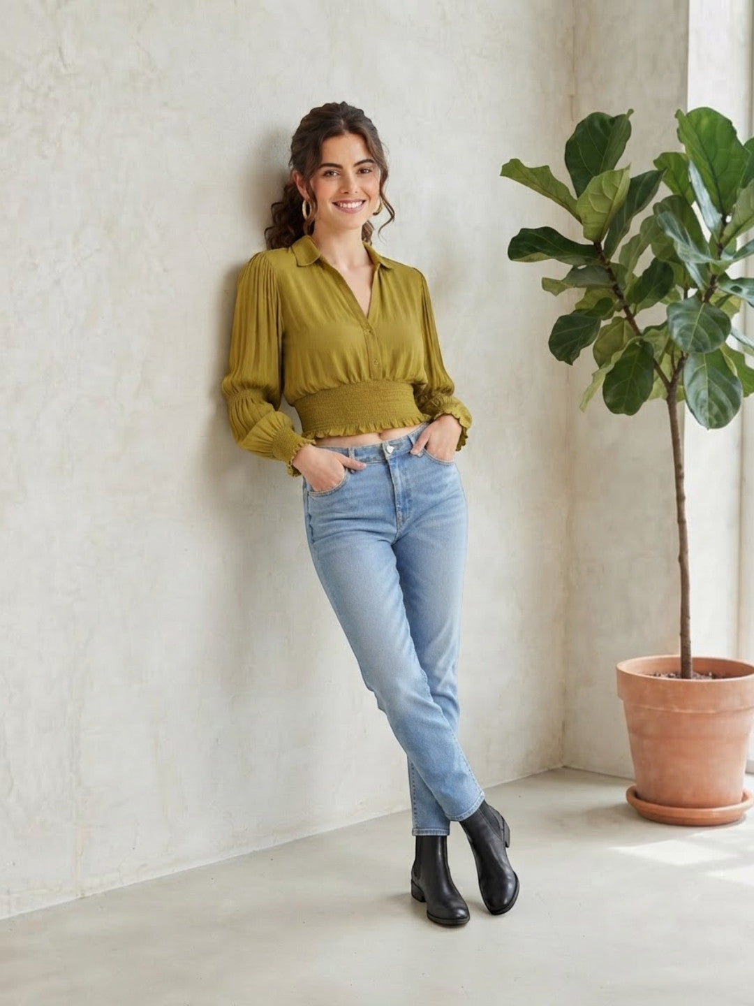Woman in olive green pleated blouse, light wash jeans, and black boots, leaning against wall near potted plant