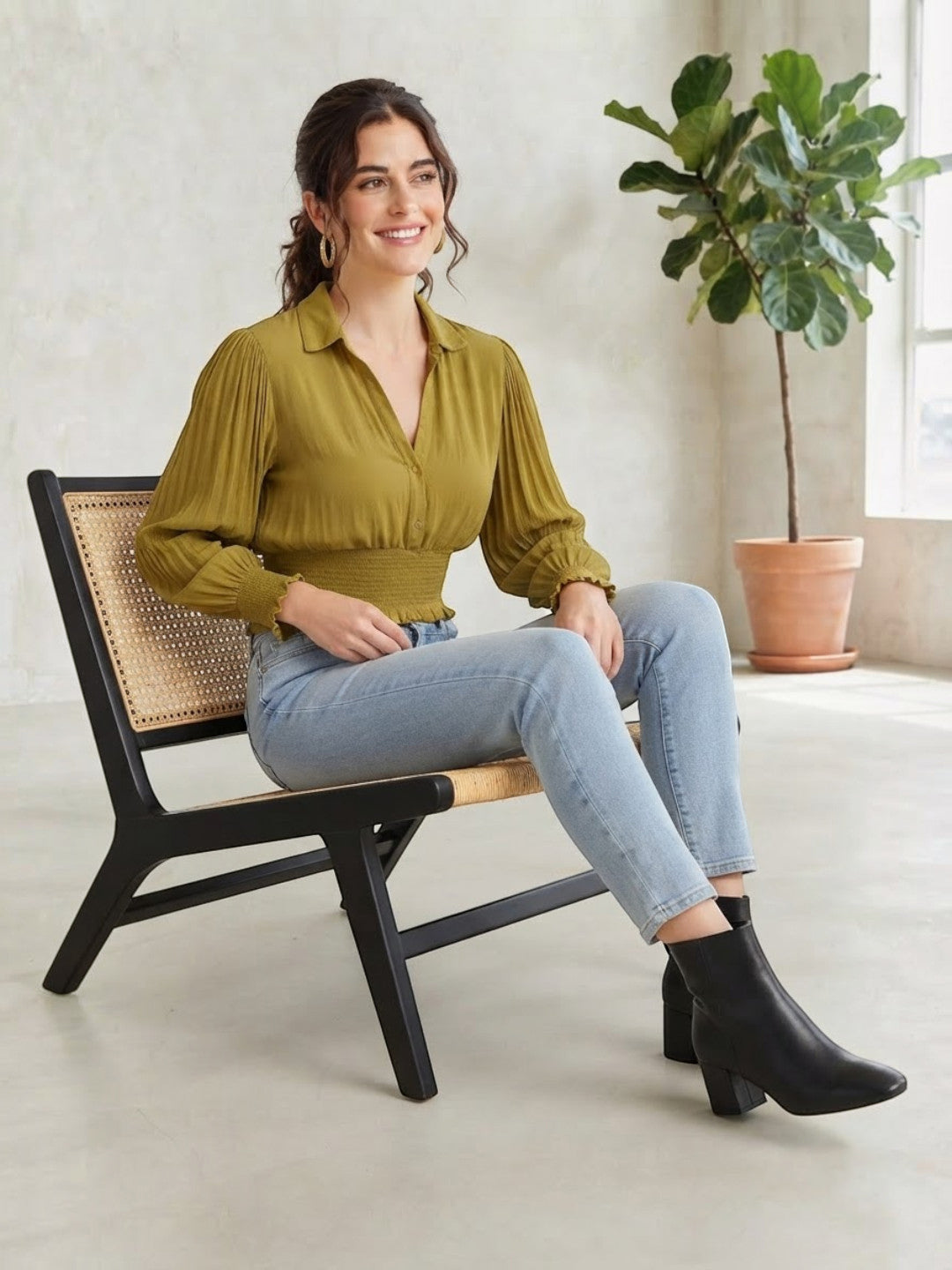 Woman in olive green pleated blouse, light wash jeans, and black ankle boots, seated on cane chair