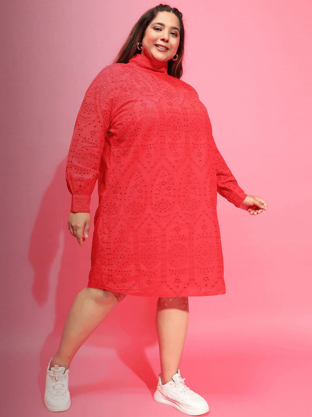 A woman wearing a red dress and sunglasses, holding a black purse, and wearing white sneakers.