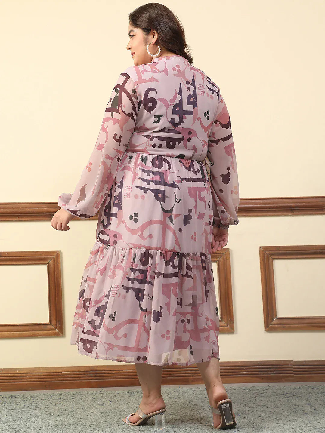 A woman wearing a pink and purple patterned dress with a high neckline and long sleeves, smiling and posing with her hand in her pocket.