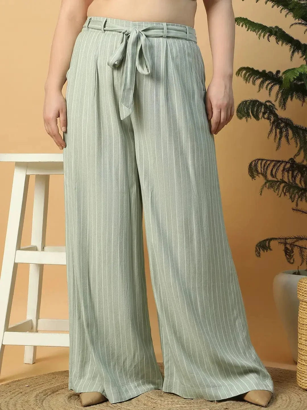 A woman wearing a white top and green striped pants is posing with her hand on her head.