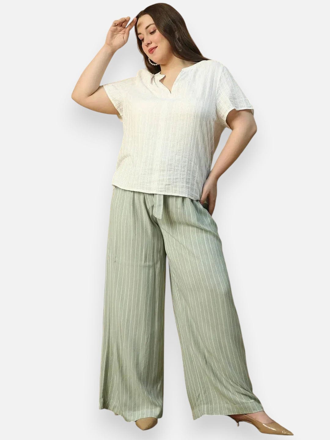 A woman wearing a white top and green striped pants is posing with her hand on her head.
