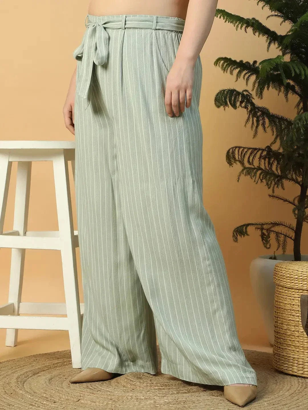 A woman wearing a white top and green striped pants is posing with her hand on her head.