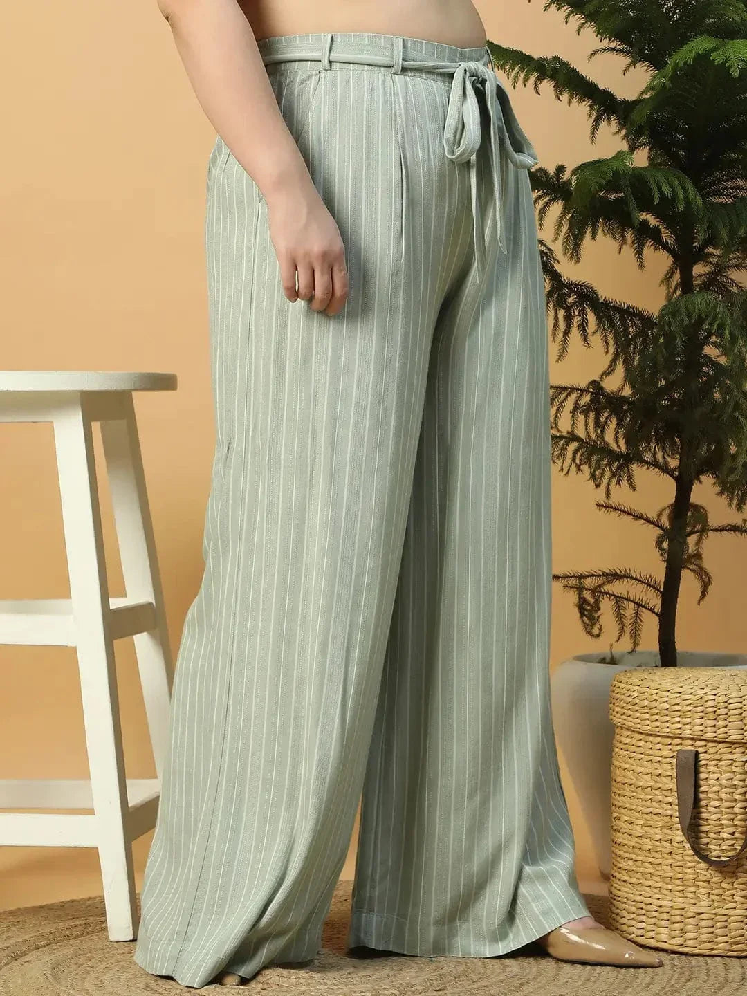 A woman wearing a white top and green striped pants is posing with her hand on her head.