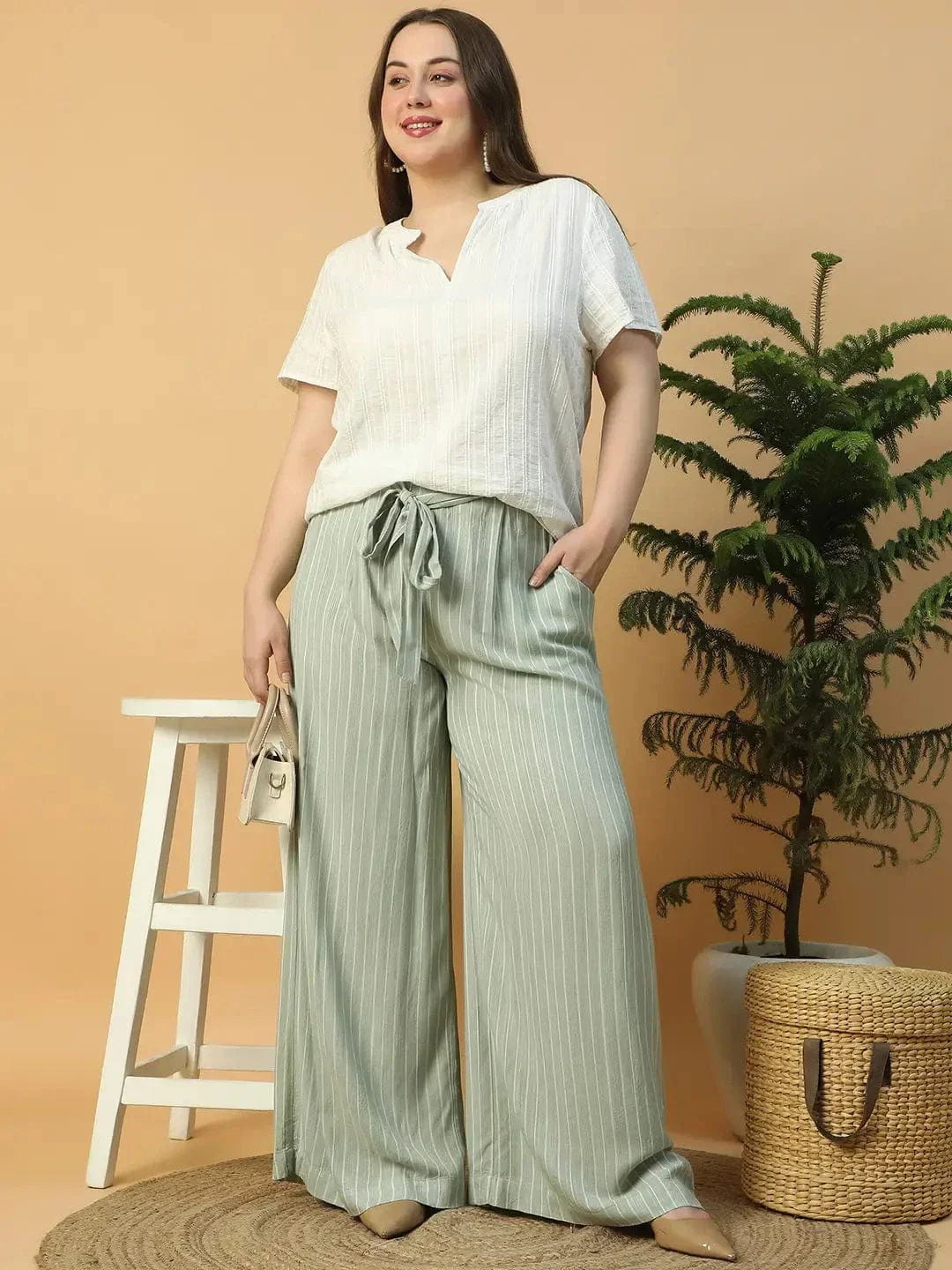 A woman wearing a white top and green striped pants is posing with her hand on her head.