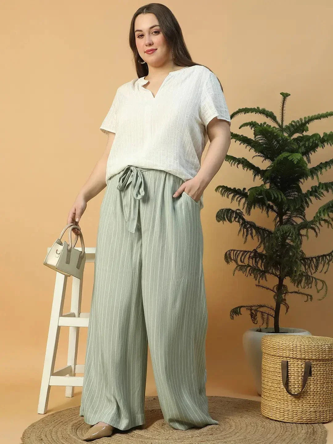 A woman wearing a white top and green striped pants is posing with her hand on her head.