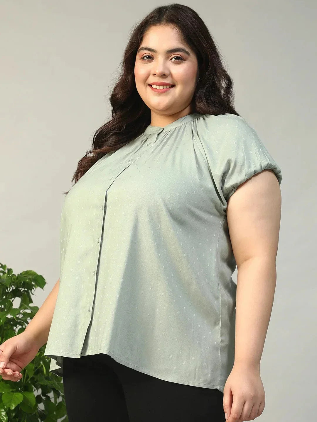 A woman with long brown hair is smiling and posing for the camera. She is wearing a light green blouse with a ruched collar and buttons.