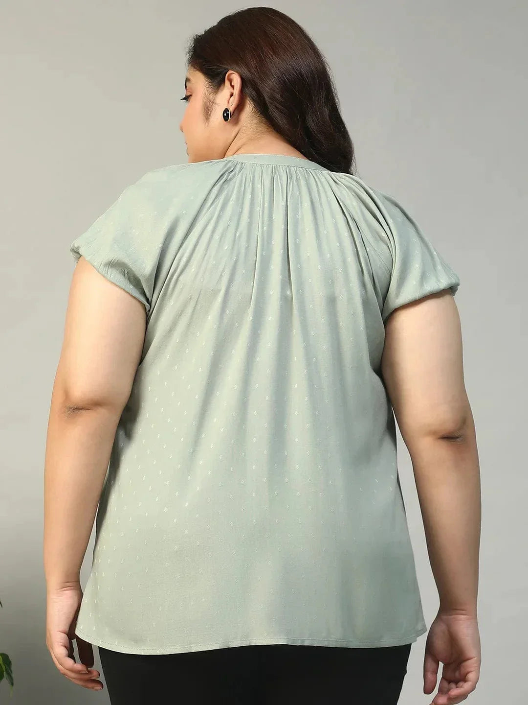 A woman with long brown hair is smiling and posing for the camera. She is wearing a light green blouse with a ruched collar and buttons.