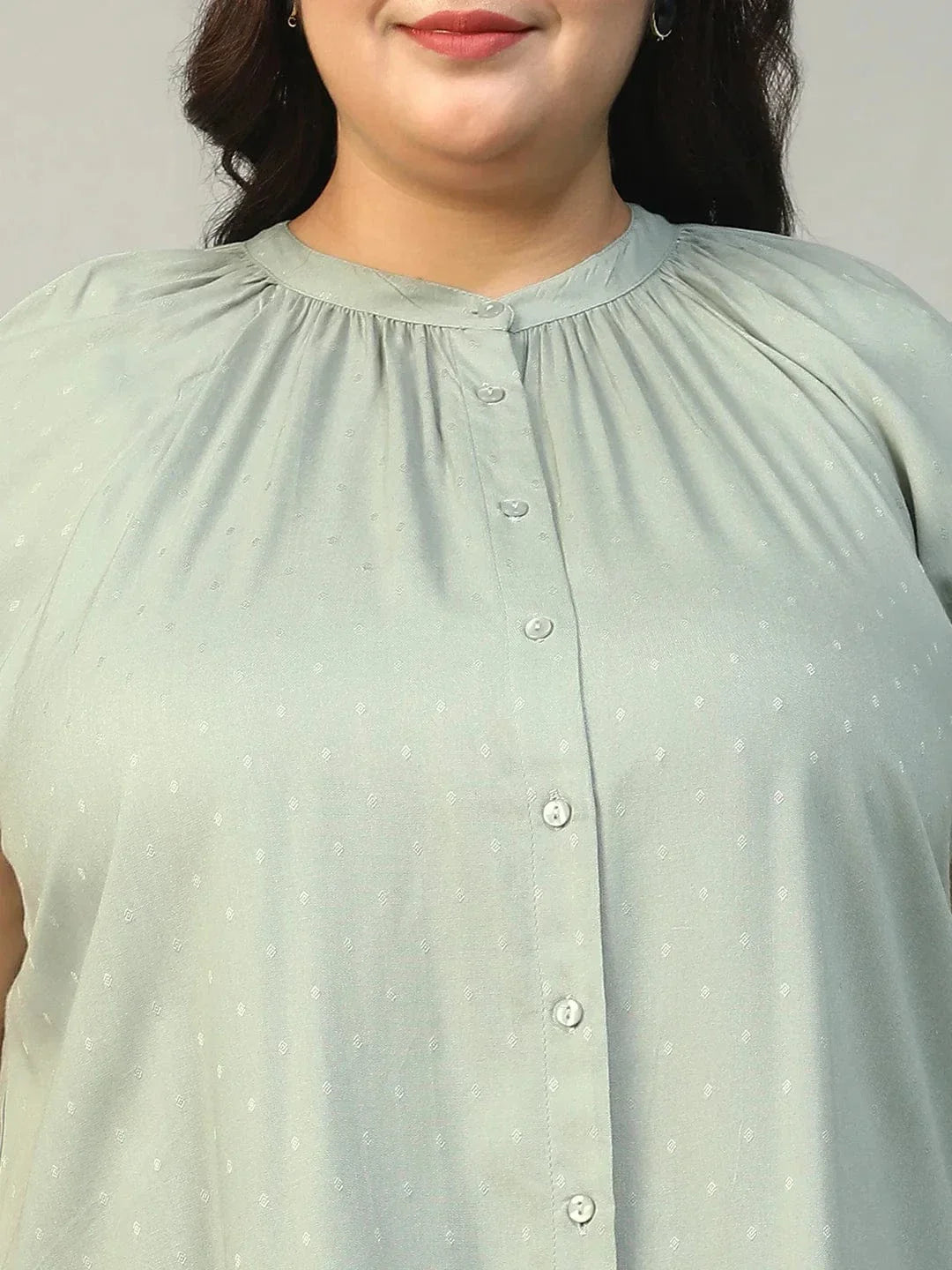 A woman with long brown hair is smiling and posing for the camera. She is wearing a light green blouse with a ruched collar and buttons.