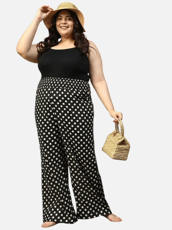 A woman wearing a black tank top, black and white polka dot pants, and a straw hat is smiling and holding a small woven bag.