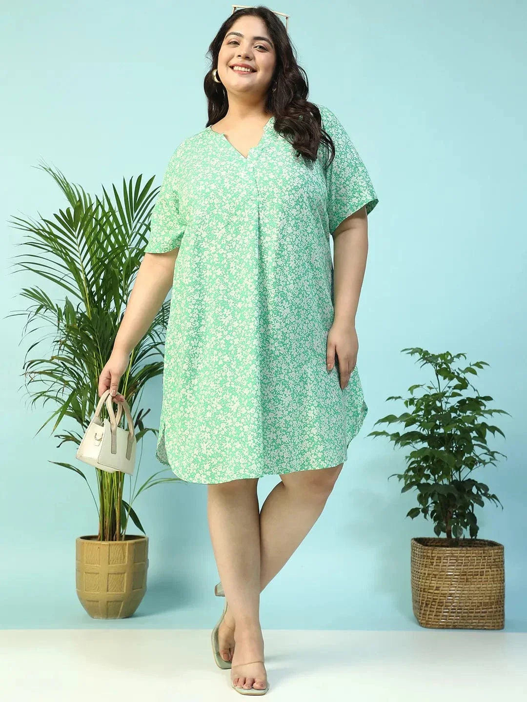 A smiling woman wearing a green floral print dress and holding a small white purse.