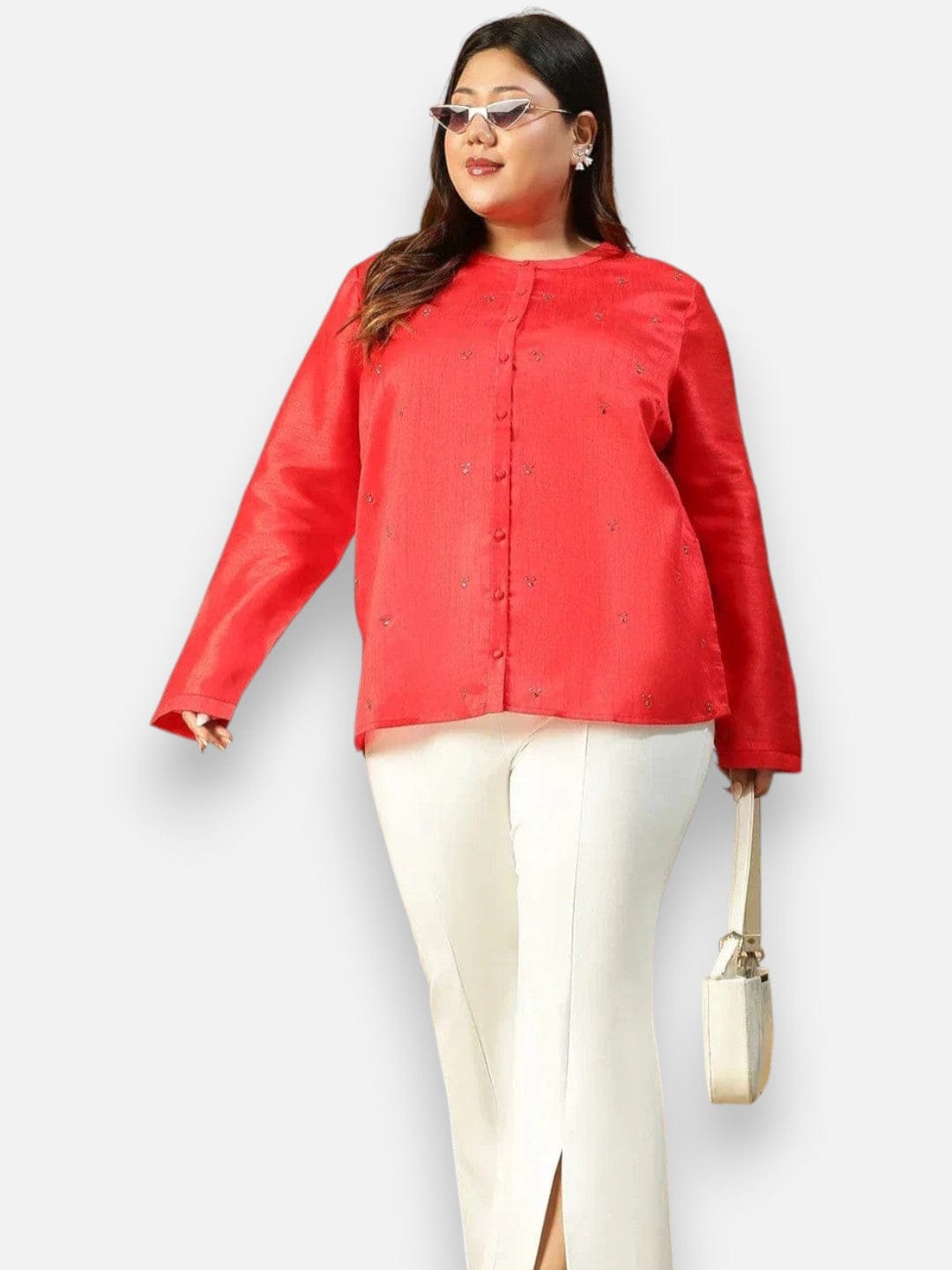 A woman wearing a red blouse with buttons and white pants, standing against a white background. She is holding a white purse in her hand.