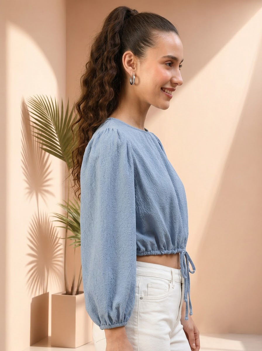 Woman in blue textured crop top with puff sleeves, white jeans, side profile, indoor plant background