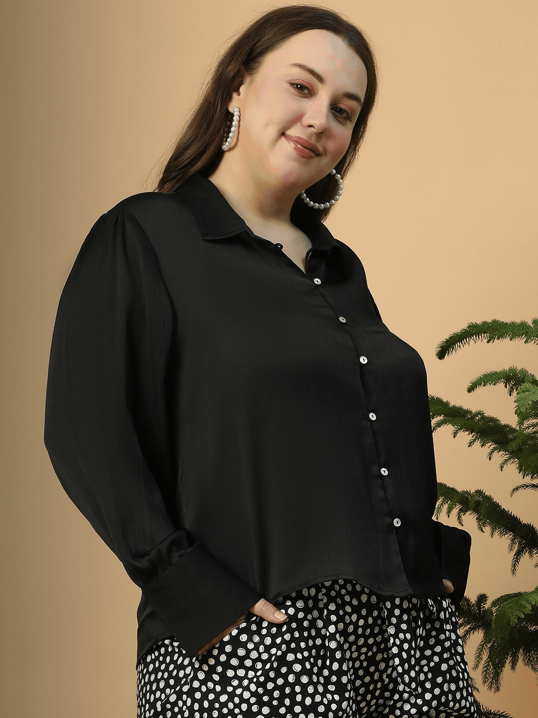 A woman wearing a black blouse and black and white patterned pants, with large hoop earrings and sunglasses, posing for a portrait against a white background.