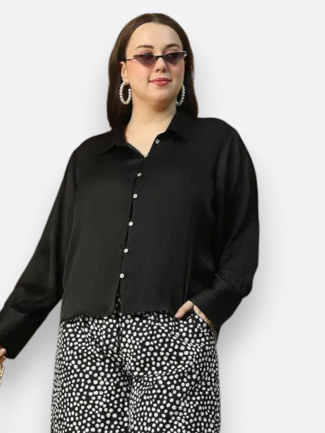 A woman wearing a black blouse and black and white patterned pants, with large hoop earrings and sunglasses, posing for a portrait against a white background.